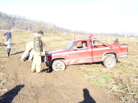 Red Dodge Dakota pickup stuck in mud with several people pushing it in a brown field