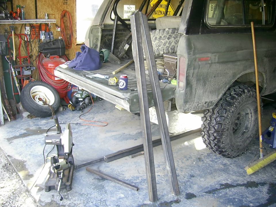Ford Bronco in garage, tailgate down, with metal bars and tools around. Spare tire and air compressor visible