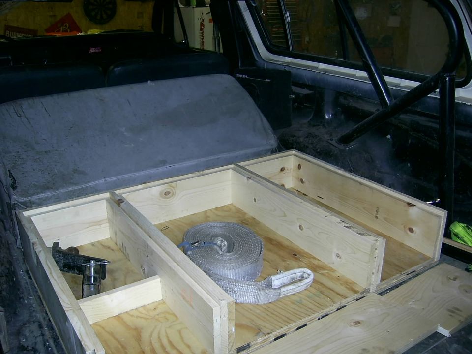 Vehicle interior with custom wooden storage box and gray seat. A rolled strap is in the box