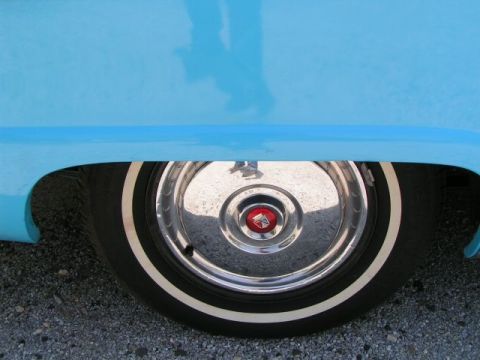 1956 Ford Fairlane - A close-up of a blue 1956 Ford Fairlane's rear wheel, a chrome hubcap with a red emblem and a white sidewall tire.