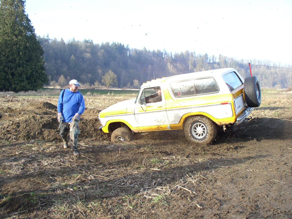 White and gold Ford Bronco with spare on rear bumper stuck in mud, a man walking beside it
