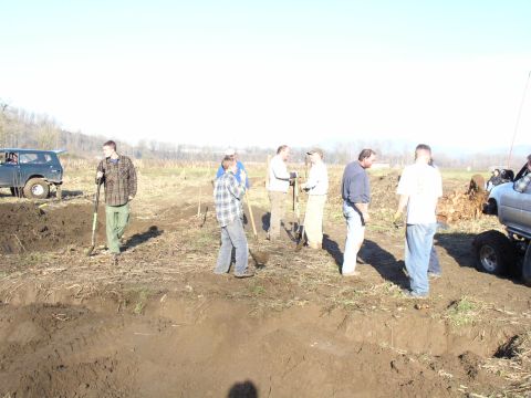 Line of men with shovels working a dirt trench in a field with trucks parked at the edges