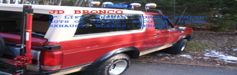 1989 Ford Bronco - A red 1989 Ford Bronco is viewed from the rear quarter, equipped with off-road accessories and a visible Hi-Lift jack.