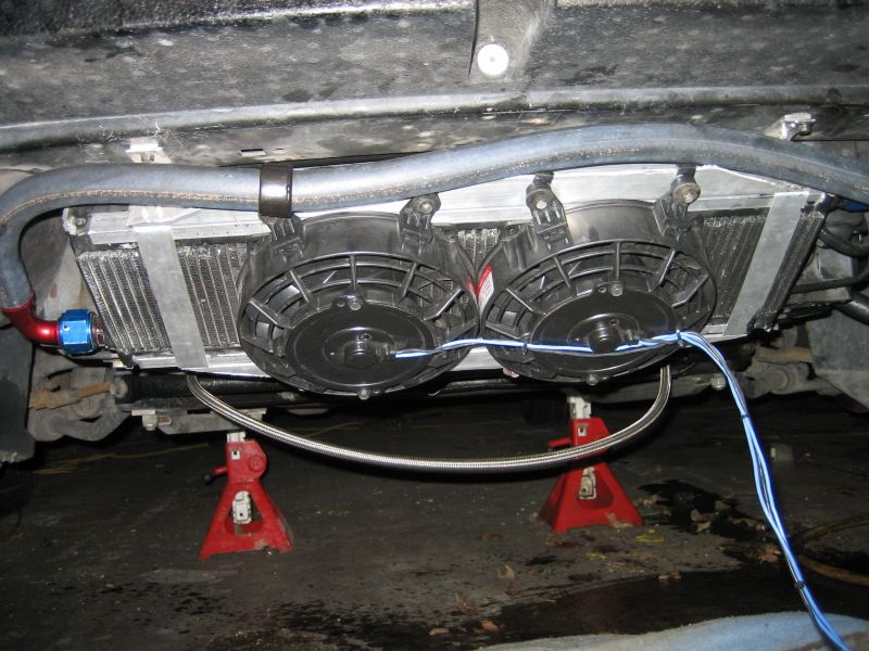 Underside of a car showing an aftermarket oil cooler with two electric fans mounted below it. The car is supported by red jack stands