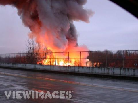 1971 Ford Ranchero - A massive fire erupts, sending thick smoke into the overcast sky, viewed from a wet road.