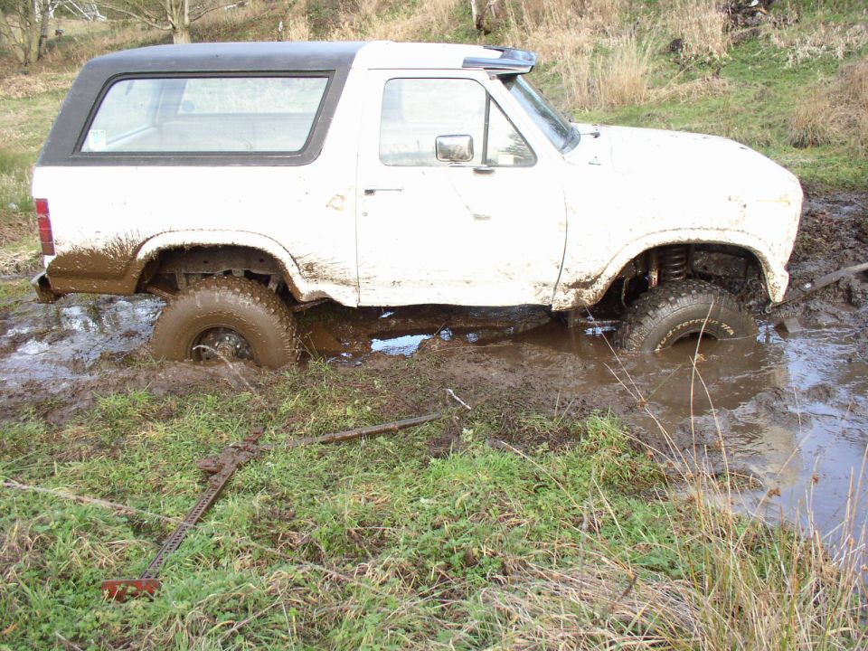 White Ford Bronco with black hardtop sunk to the axles in a muddy puddle in a field