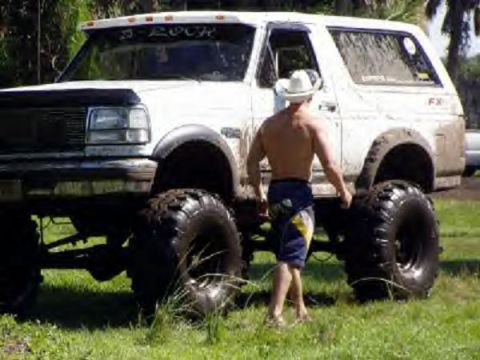 1995 Ford Bronco - A man in a cowboy hat stands beside a heavily modified white 1995 Ford Bronco, its massive off-road tires.