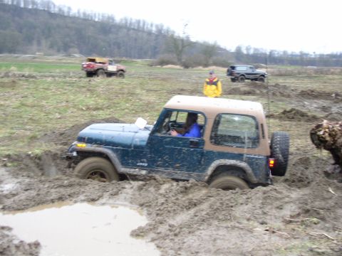 Blue Jeep Wrangler with tan top sunk in a deep mud pit, other trucks and a spotter behind