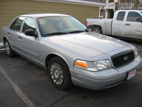 2004 Ford Crown Victoria - Silver sedan with a black grille and chrome wheel covers is parked in front of a building, with a pickup truck behind it.