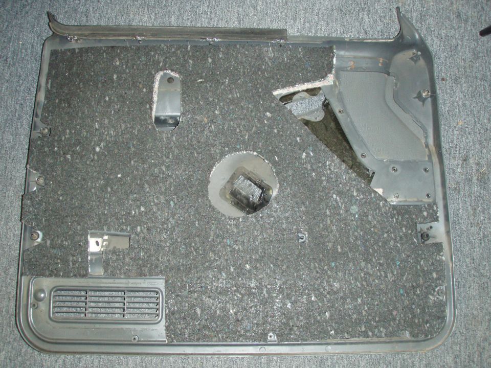 Back of a gray door panel with fiber insulation, brackets, and vent grille, lying on carpet