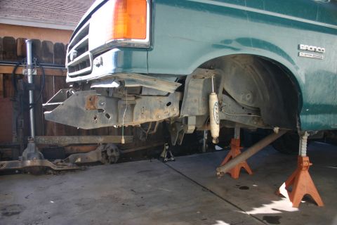 1991 Ford Bronco - Front suspension and frame of a green 1991 Ford Bronco are visible while it is elevated on jack stands.