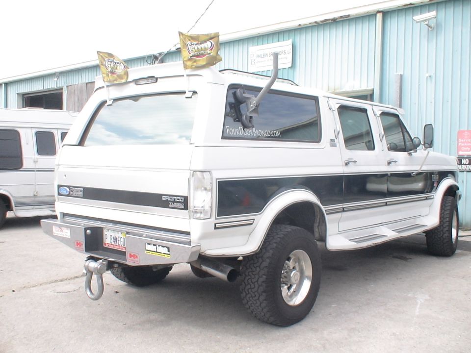 White Ford Bronco SUV with dark stripe, aftermarket bumper, and two flags on roof. Another van is parked to the left