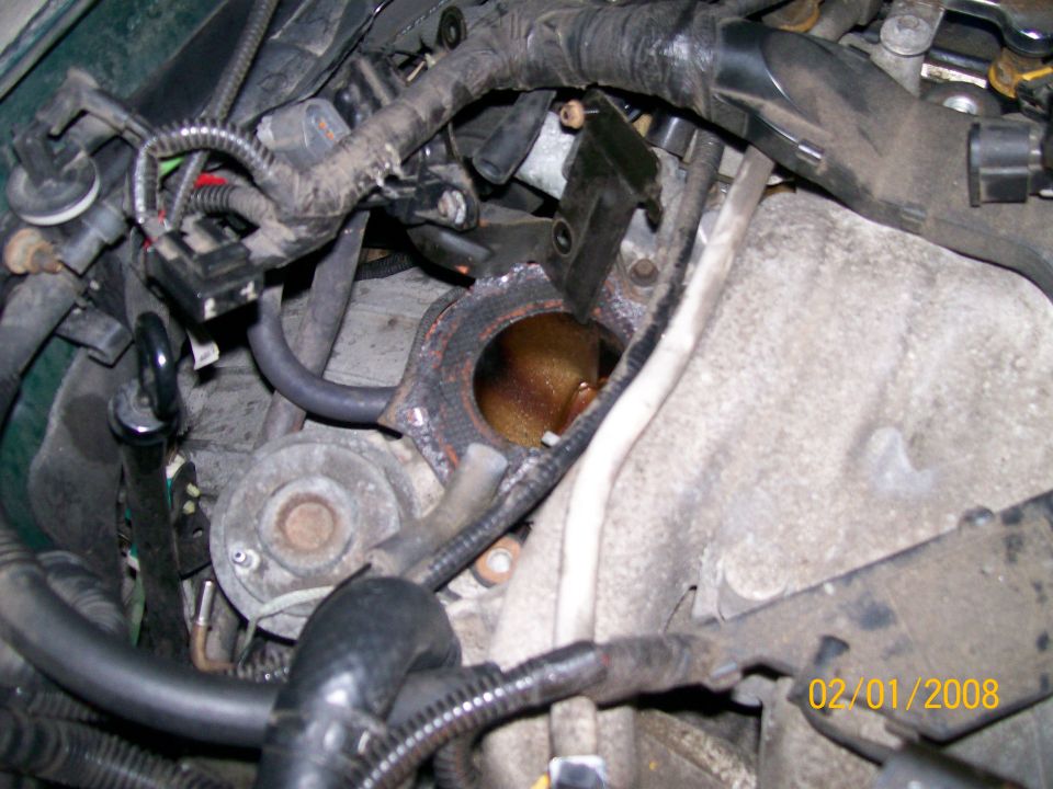 Engine bay close-up of exposed exhaust manifold flange amid wiring harness and hoses