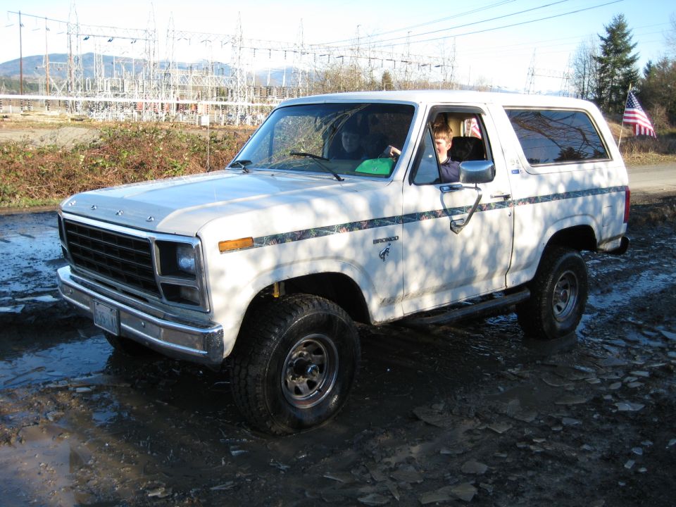 White Ford Bronco with camouflage stripe, parked in mud. Visible grille, headlights, and side profile