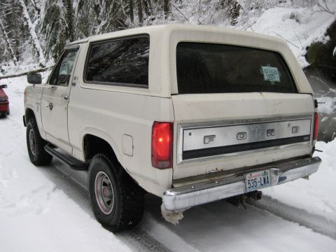 1980 Ford Bronco - A white 1980 Ford Bronco drives through snow-covered terrain, with a red vehicle partially visible on the left.