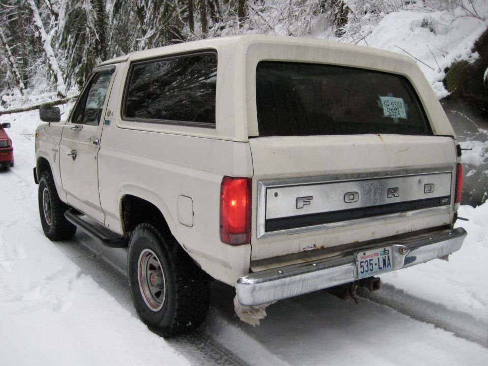 White 1980 Ford Bronco in snow, rear view, with a red car visible to the left