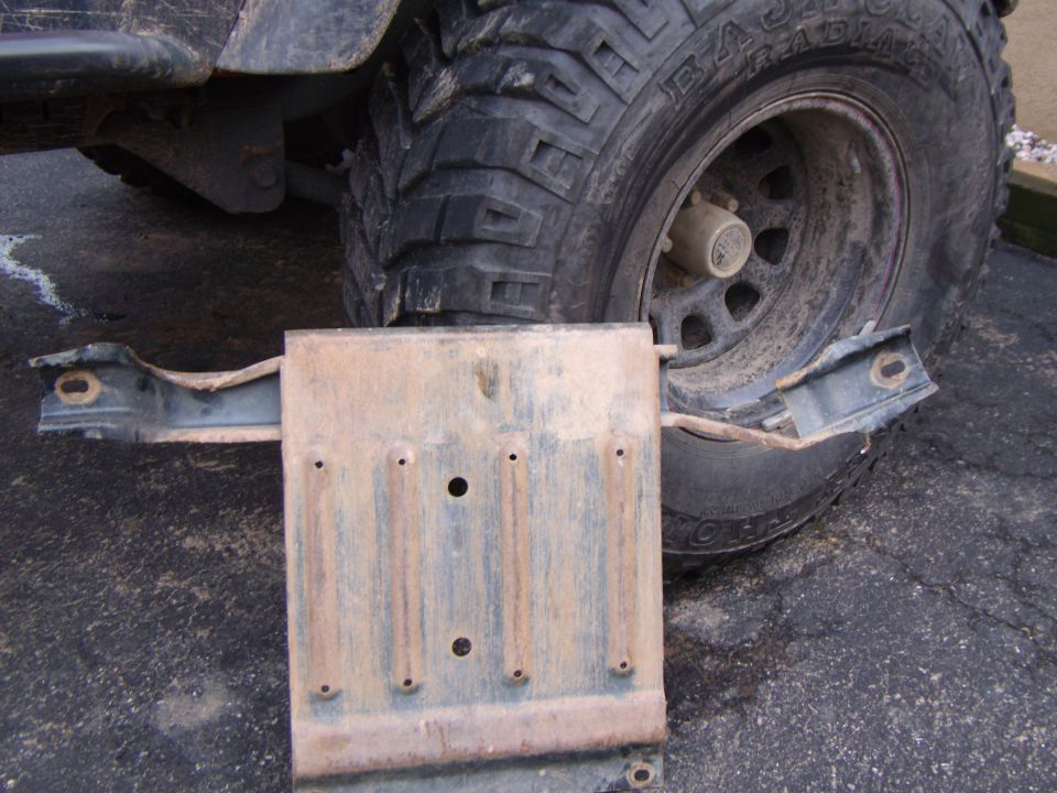 Rusty metal transmission pan skid plate with mounting brackets, next to a large off-road tire