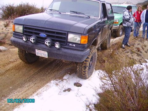 1990 Ford Bronco - A dark gray Ford Bronco navigates a muddy and snowy off-road trail, with other vehicles and people visible in the background.
