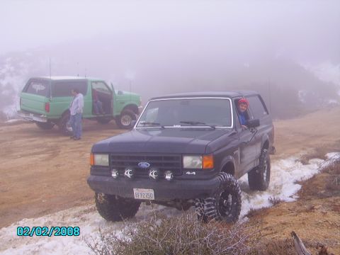 1990 Ford Bronco - Two Ford Broncos navigate a foggy, snowy dirt road, likely off-roading.