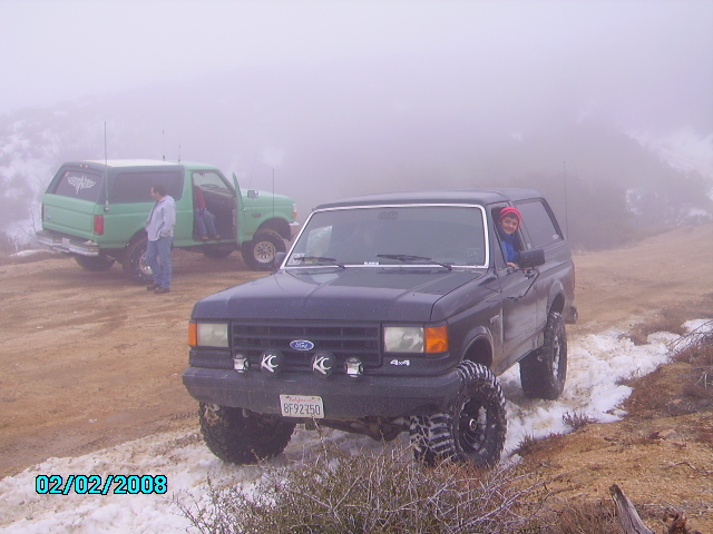 1990 Ford Bronco in front, another green Bronco behind it, driving on a dirt road with snow patches