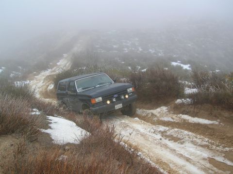 1985 Ford Bronco - A Ford Bronco navigates a challenging off-road path with patches of snow and mud under a foggy sky.