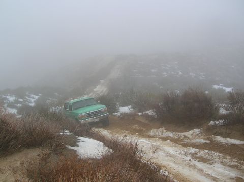 1985 Ford Bronco - A green pickup truck navigates a challenging off-road path obscured by heavy fog and patches of snow.