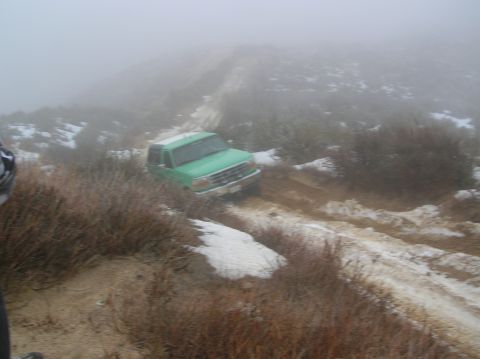 1985 Ford Bronco - A green SUV navigates a snowy and muddy dirt road in foggy conditions.