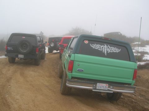 1985 Ford Bronco - A convoy of Ford Broncos navigates a dusty, off-road trail shrouded in fog.