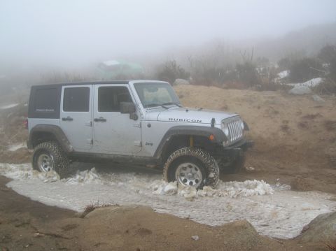 1985 Ford Bronco - A Jeep Wrangler navigates a muddy and slushy terrain, with a second vehicle obscured by fog in the distance.