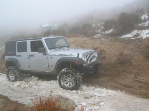 1985 Ford Bronco - A silver Jeep Wrangler Rubicon navigates a snowy and muddy trail, with another vehicle visible in the foggy background.