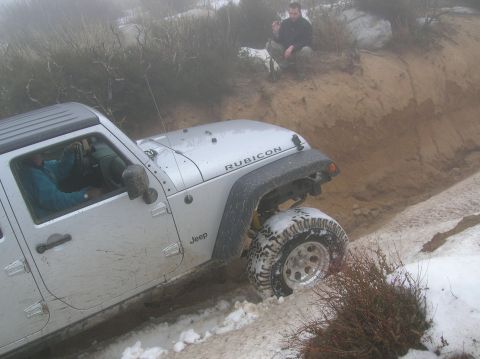 1985 Ford Bronco - A Jeep Wrangler navigates a snowy and muddy trail with a passenger observing from higher ground.