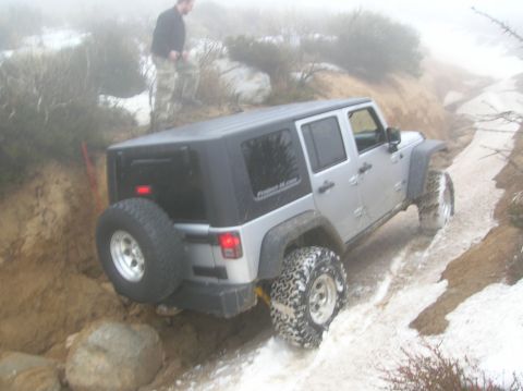 1985 Ford Bronco - A silver Jeep Wrangler navigates a challenging snowy and muddy off-road trail with a person observing from a nearby incline.
