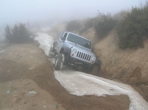 1985 Ford Bronco - A silver Jeep Wrangler navigates a muddy and snowy trail in foggy conditions.