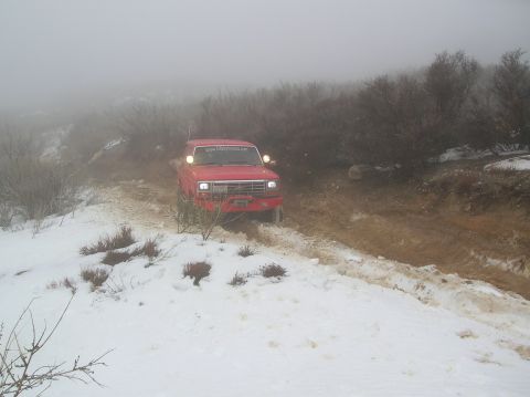 1985 Ford Bronco - A red pickup truck navigates a challenging off-road path covered in snow and mud.