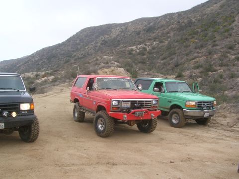 1985 Ford Bronco - Three modified Ford Broncos are parked on a dirt road in a hilly, arid landscape.