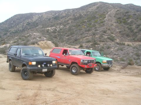 1985 Ford Bronco - A trio of Ford Bronco SUVs are parked on a dirt trail, with a mountainous, arid landscape in the background.