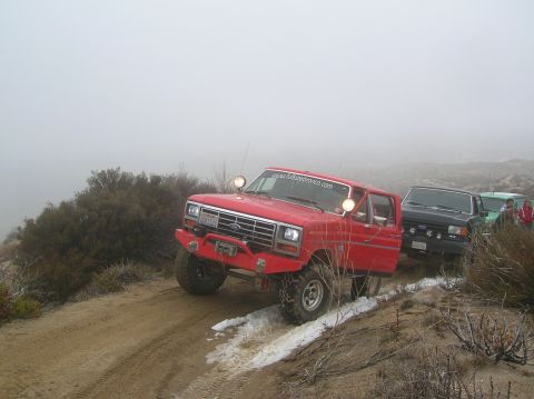 1985 Ford Bronco - A red 1985 Ford Bronco navigates a foggy, off-road trail with other vehicles following.