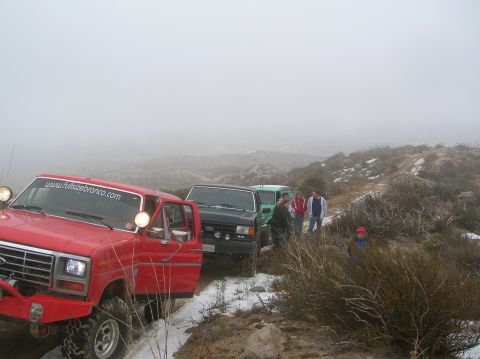 1985 Ford Bronco - A line of off-road vehicles, including a red Ford Bronco, navigate a foggy, mountainous terrain.