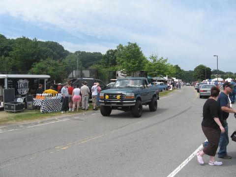1996 Ford Bronco - A lifted green pickup truck drives past a roadside event with people and vendors.