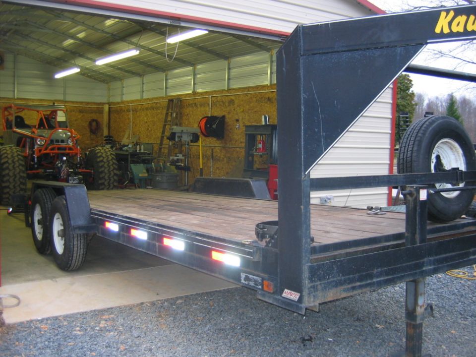 Flatbed trailer parked in garage with a large off-road vehicle on it. Spare tire mounted on trailer
