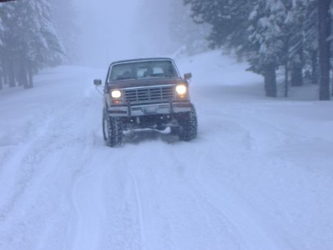 1986 Ford Bronco - A red SUV navigates a snow-covered road, its headlights cutting through the wintery conditions.