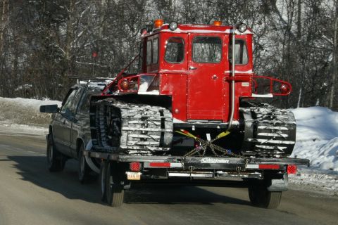 2003 Jeep TJ - A truck transports a red tracked vehicle, likely a snow groomer, on a flatbed trailer during winter.