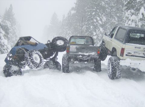 1986 Ford Bronco - Several off-road vehicles are stuck in deep snow on a wooded trail during a snowstorm.