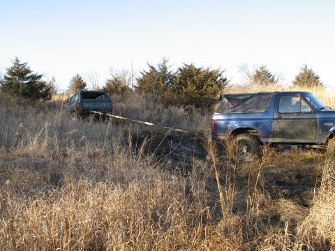 1995 Ford Bronco - A blue SUV is being towed through a field of dry grass by another vehicle.