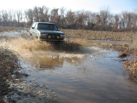 1996 Ford Bronco - A green Ford Bronco navigates a flooded dirt road, spraying mud and water.