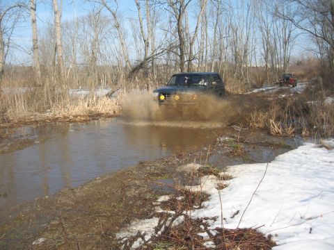 1996 Ford Bronco - A 1996 Ford Bronco navigates a muddy obstacle, kicking up a large spray of water.