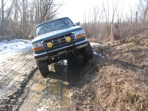1996 Ford Bronco - Green 1996 Ford Bronco navigates a muddy, off-road trail with snow patches visible.