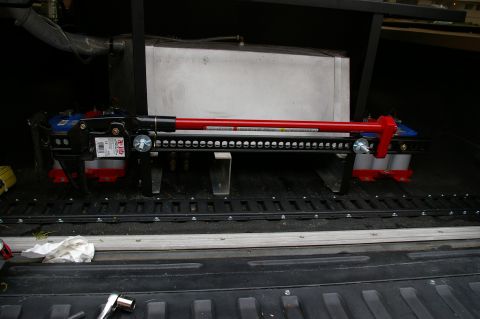 1986 Ford Bronco - A Hi-Lift jack is stored in the bed of a pickup truck, resting on tie-down rails.