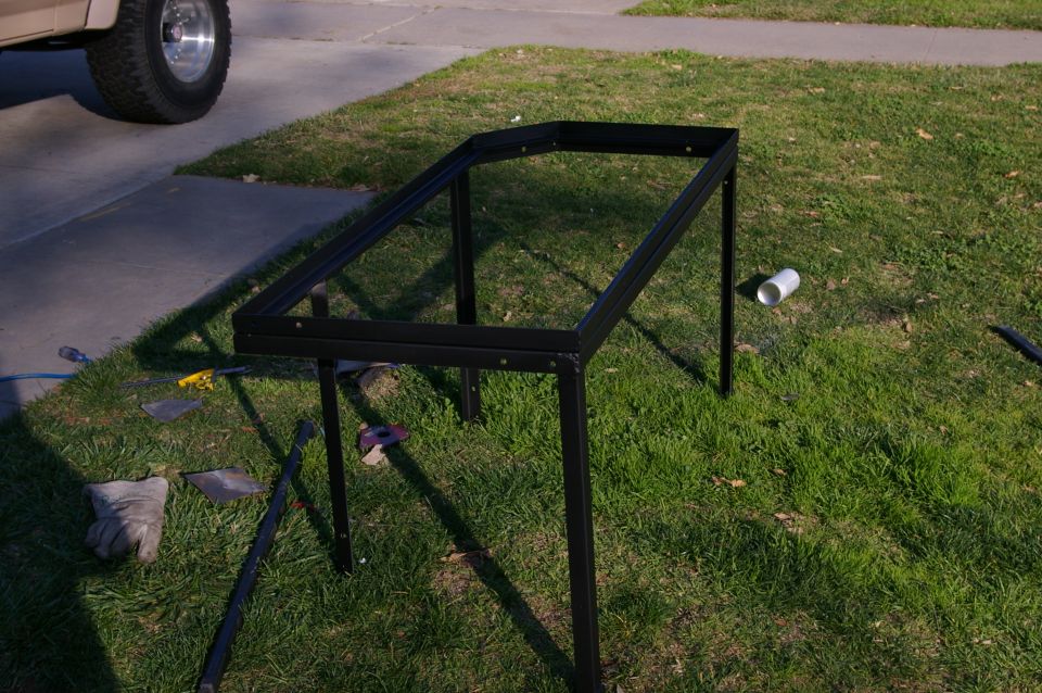 Black metal frame with four legs sitting on grass next to a sidewalk. A truck tire is visible in the upper left corner