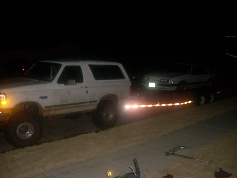 1992 Ford Bronco - Night shot of a mud-splattered lifted white full-size Ford Bronco with hardtop, side view, hitched to a dual-axle trailer loaded with a whi…